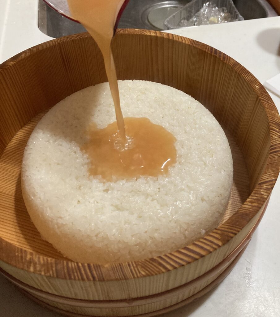 炊きたてのご飯に柿酢をかけて酢飯を作る様子。木の飯台に入ったご飯に酢を注いでいる。