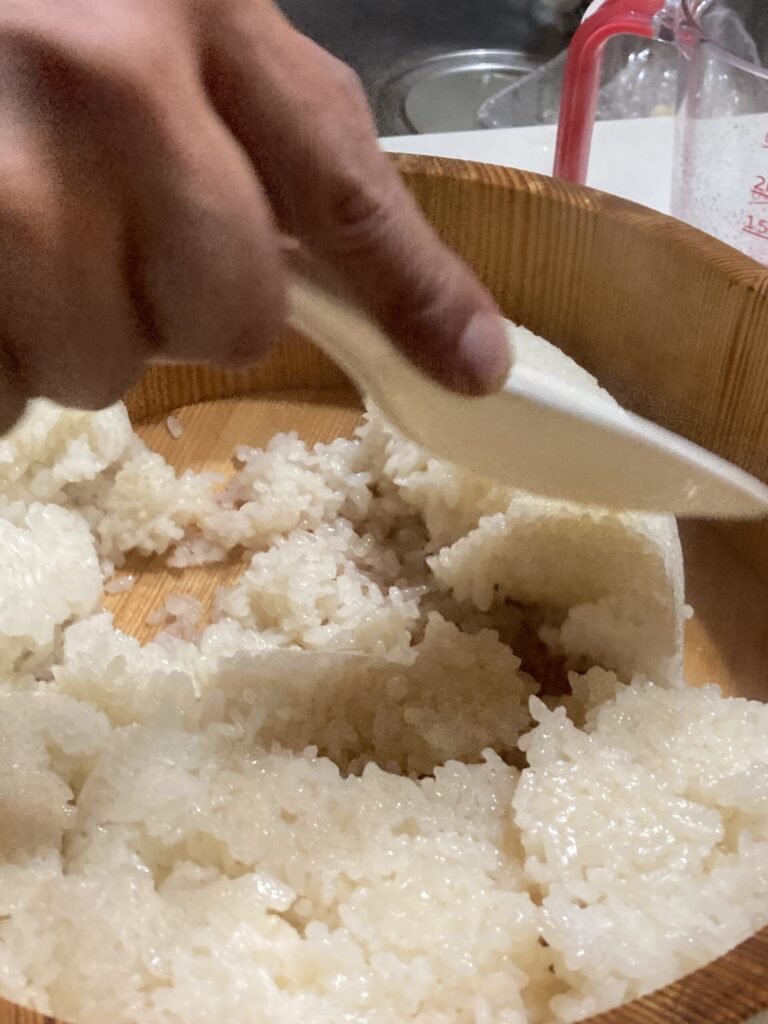 **alt（代替テキスト）**
しゃもじで切るように混ぜて酢飯を作っている様子。飯台の中でご飯を縦切りに混ぜている。
